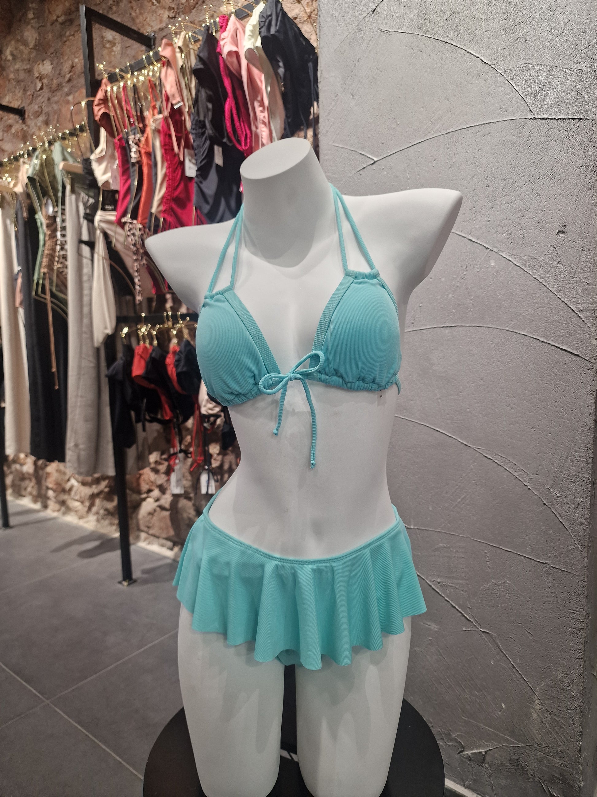 Turquoise bikini top on a mannequin bust with white ruffled garment against a gray tiled floor.