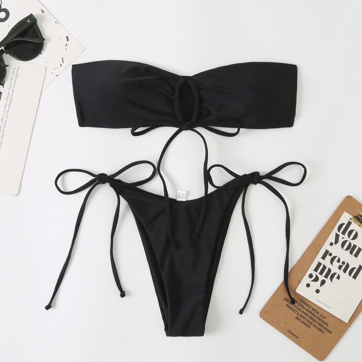 Black bikini set on a white surface with sunglasses and a card in the background

